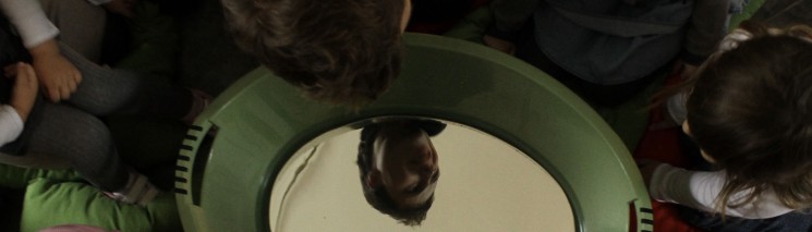 Dzieci siedzące w kółku nachylają się nad dużym okrągłym lustrem. 