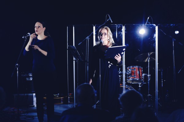 Dwie kobiety stoją na scenie, jedna przy mikrofonie czyta z kartki. 
