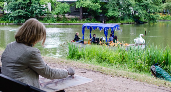 Kobieta siedzi w parku i maluje, przed nią znajduje się rzeka, po której płynie gondola.