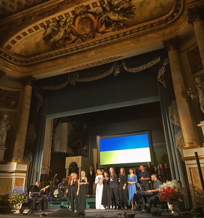 Grupa artystów stoi na scenie Teatru Królewskiego i śpiewa. Nad artystami wisi ukraińska flaga w niebiesko-żółtych barwach.