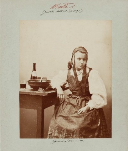 Dawna fotografia ukazująca dziewczynę w stroju ludowym siedzącą przy stole, na którym stoi miska, butelka i szklanka.