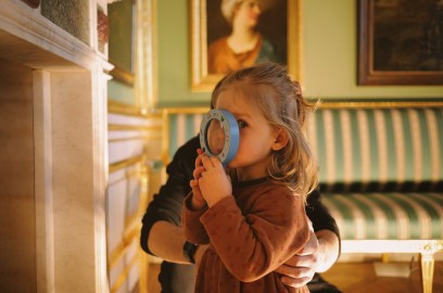 Mała dziewczynka patrząca przez lupę, którą trzyma w dłoni. Dziewczyna stoi w muzealnej sali, w której na ścianach wiszą obrazy i stoi kanapa w biało-zielone pasy. 