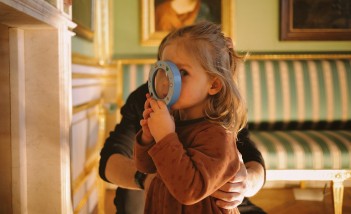 Mała dziewczynka patrząca przez lupę, którą trzyma w dłoni. Dziewczyna stoi w muzealnej sali, w której na ścianach wiszą obrazy i stoi kanapa w biało-zielone pasy. 