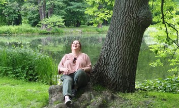 Kobieta siedząca w parku pod drzewem przy stawie.