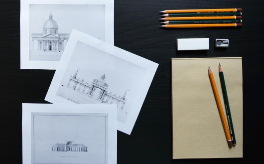 Rysunki przedstawiające budynki oraz kredki i ołówki leżące na blacie, dwa z ołówków leżą na kartce papieru. 