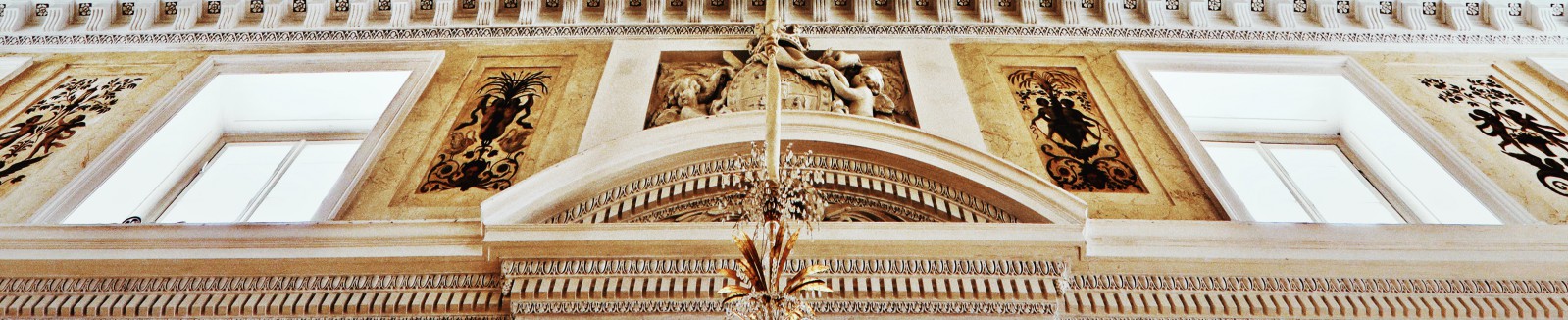 Fragment ściany w Sali Balowej w Pałacu na Wyspie. Ściana ozdobiona jest poziomymi sztukateriami. Znajdują się na niej dwa oka, między oknami są płaskorzeźby przedstawiające amorki, przy oknach ścianę zdobią malowidła z motywami roślinnymi i postaciami. 