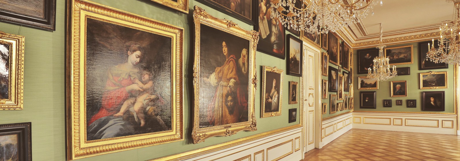 Galeria Obrazów w Pałacu na Wyspie. Obrazy z kolekcji króla Stanisława Augusta w złotych ramach wiszą na jasnozielonej ścianie. Z sufitu zwisają trzy kryształowe żyrandole.