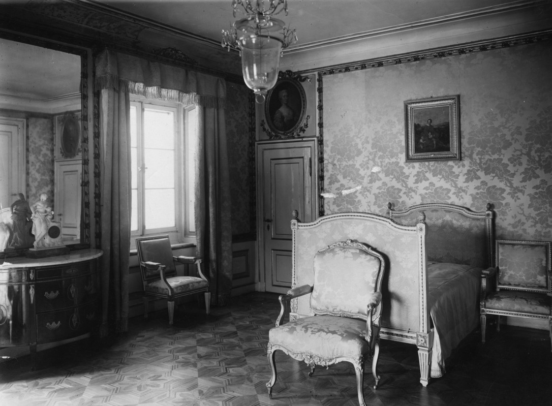 Sypialnia królewska w Pałacu na Wyspie. Pośrodku stoi łóżko, przy nimi krzesła, po lewej stronie stoi komoda, dalej jest okno z zasłonami, pod którym stoi krzesło. 