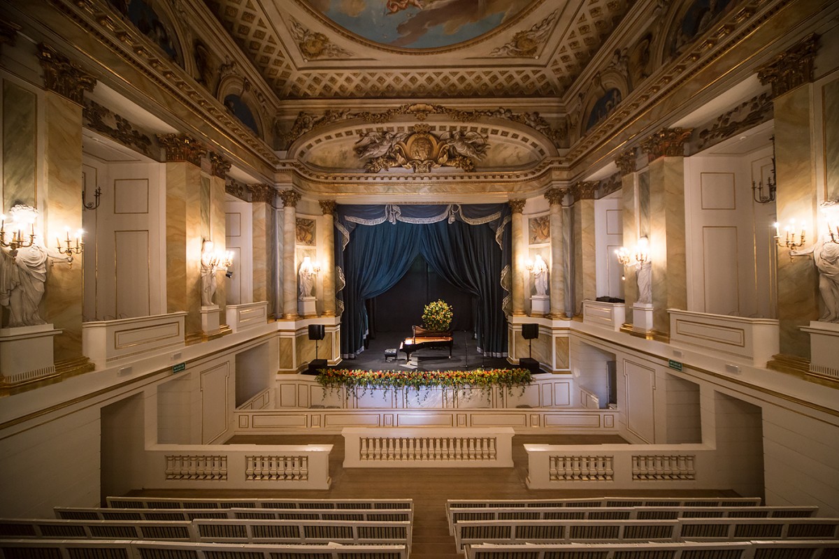 Wnętrze Teatru Królewskiego w Starej Oranżerii, na scenie ozdobionej kotarą stoi fortepian, pod ścianami znajdują się rzeźby trzymające w rękach świeczniki.