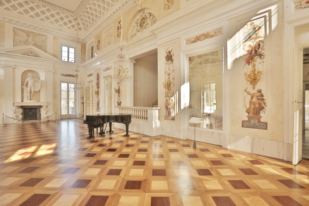 Sala Balowa w Pałacu na Wyspie, której ściany zdobią malowidła. W pomieszczeniu stoi fortepian. Pod ścianą przy oknie znajduje się kominek, na którym stoi posąg przedstawiający mężczyznę.