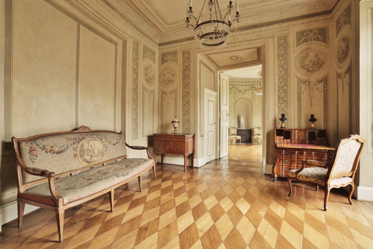 Sypialnia w Pałacu Myślewickim, po lewej stronie po ścianą stoi kanapa, na ścianie środkowej znajdują się otwarte drzwi, po ścianą stoją meble.