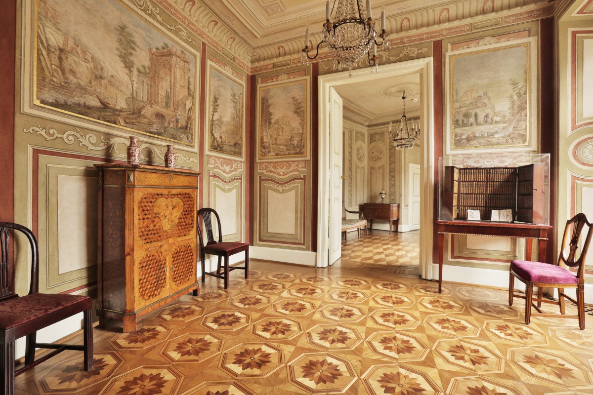 Pokój krajobrazowy w Pałacu Myślewickim, ściany zdobią malowidła przedstawiające krajobraz, z sufitu zwisa ozdobny żyrandol, po ścianami stoją komody i krzesła.