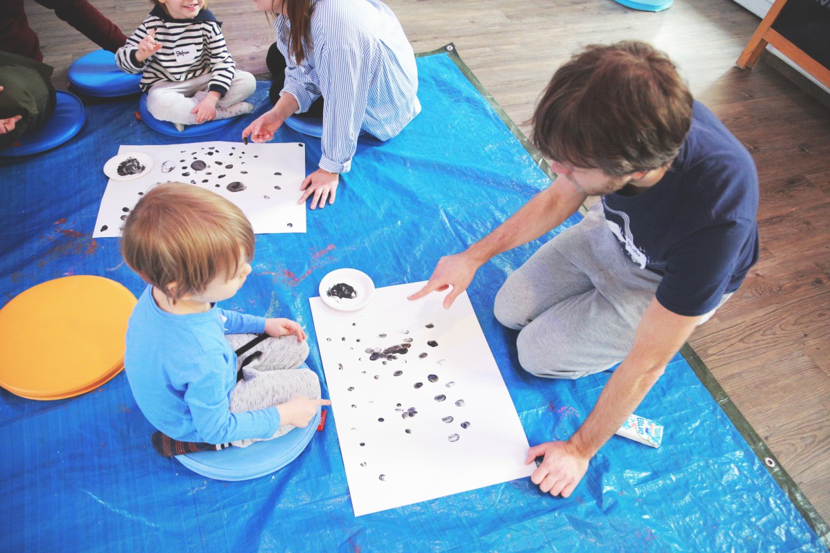 Dzieci siedzą na podłodzie i robią palcami czarne kropki na białym papierze.