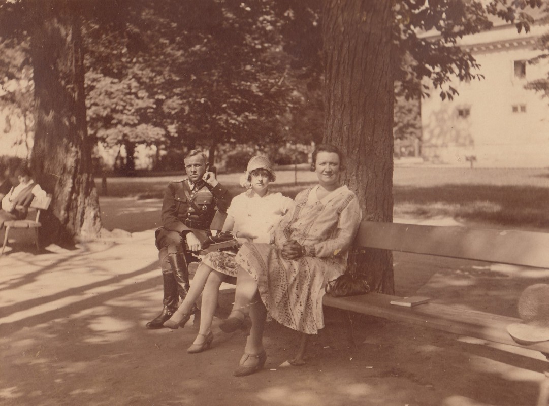 Mężczyzna w mundurze i dwie kobiety siedzą na ławce pod drzewem. 