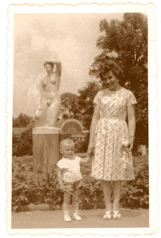 Kobieta trzyma małe dziecko za rękę, stoją w ogrodzie, z tyłu widoczny jest posąg kobiety. 