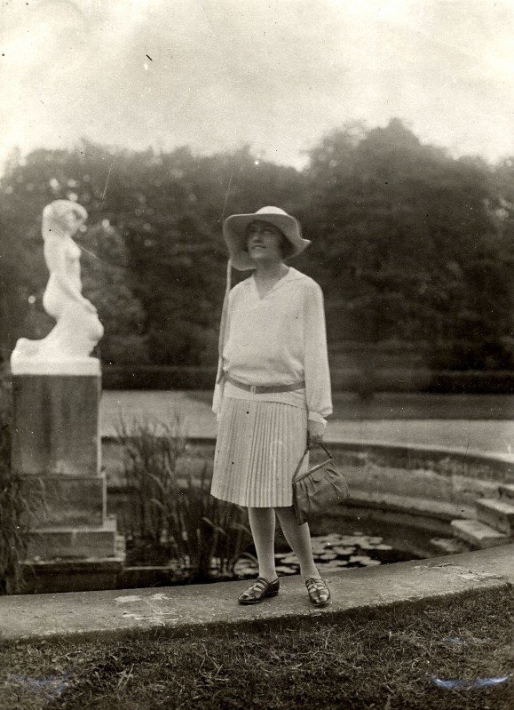 Kobieta z torebką w dłoni, stojąca przy fontannie, w tle widać rzeźbę przedstawiającą postać kobiety. 
