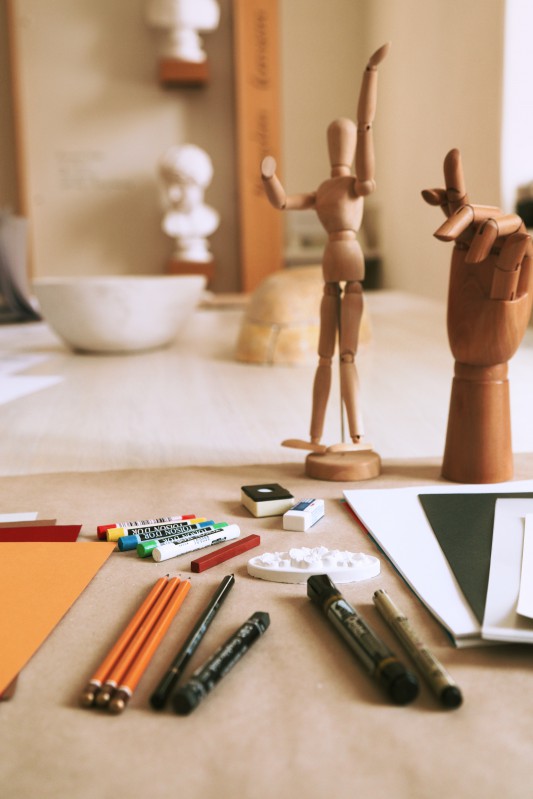 Zestaw malarski składający się z kartek kreślarskich, ołówków, kredek i gumki, obok stoi drewniane figurki przedstawiające postać i rękę ze zgiętym palcem.