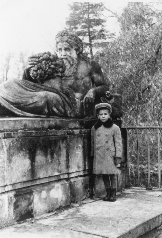 Chłopiec stojący przy rzeźbie przedstawiającej leżącego, półnagiego mężczyznę, z długimi włosami i brodą, z wieńcem laurowym na głowie i rogiem obfitości, uosabiającego rzekę Bug.