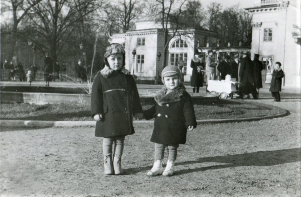 Dwóch kilkuletnich chłopców stoi na parkowej alejce, w tle widać Pałac na Wyspie i spacerujących przed budynkiem ludzi. 
