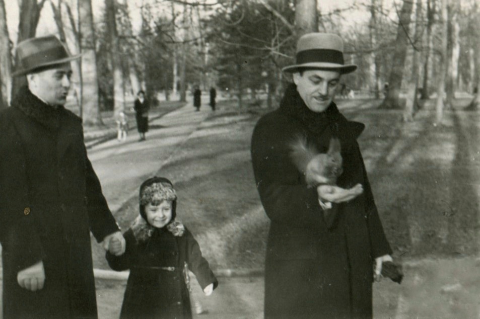Na parkowej alejce stoi mężczyzna, który trzyma za rękę kilkuletniego chłopca, obok stoi drugi mężczyzna, który ma na ręku wiewiórkę, w tle w parku spacerują ludzie.