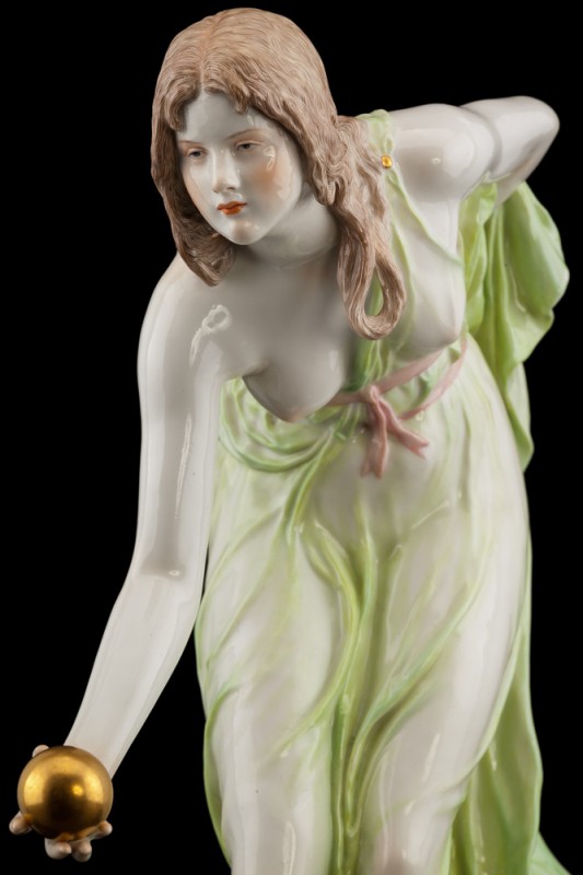 Figurka przedstawiająca młodą kobietę, pochyloną do przodu. Okrywa ją plisowana, drapowana, delikatna szata, odsłaniająca jej prawą pierś, z kokardką w talii.