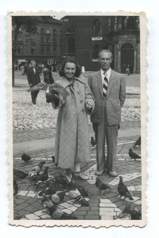 Kobieta i mężczyzna stoją na miejskim rynku. Wkół nich zebrały się ptaki. 
