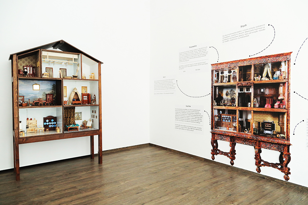 Pomieszczenie wystawowe, pod dwiema ścianami stoją plansze przedstawiające wnętrza miniaturowych domków.