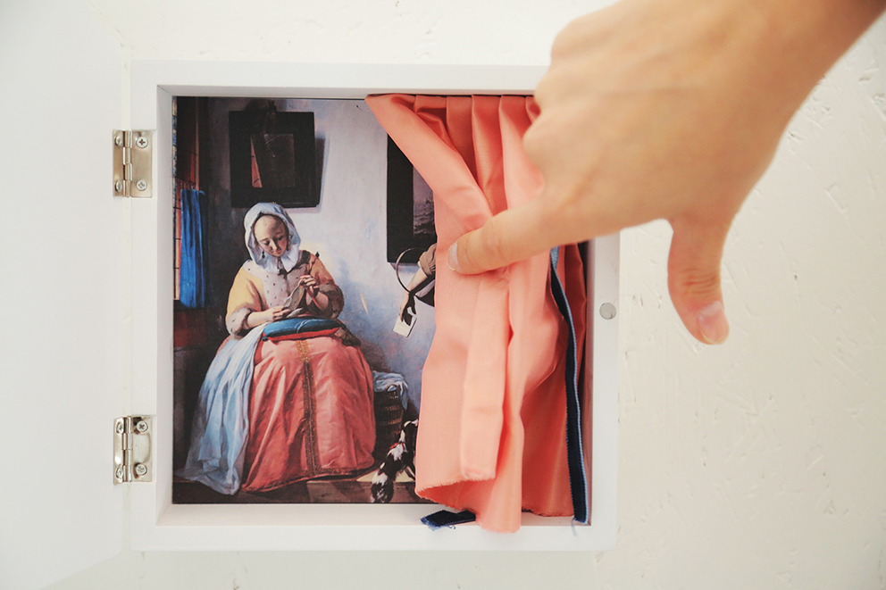 Kobieca ręka odsłania miniaturową kotarę, za którą jest obraz przedstawiający siedzącą starszą kobietę w stroju z epoki.