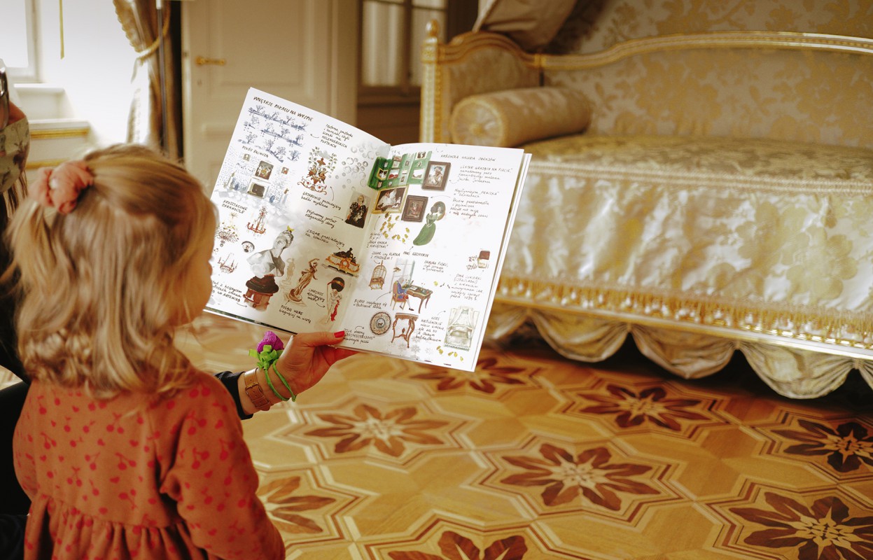 Dziewczynka trzymająca przed sobą otwartą książkę z obrazkami. Przed dziewczynką stoi zabytkowe łóżko. 