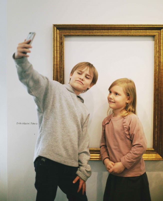 Chłopiec i dziewczynka stoją pod ścianą, na której wisi pusta rama, i robią sobie zdjęcie.