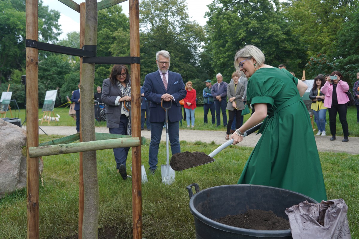 Kobieta w zielonej sukience stoi w ogrodzie z łopatą w ręku i podsypuje ziemię pod drzewo.