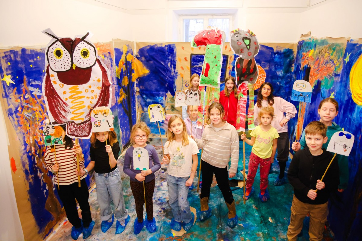 Grupa dzieci stoi na tle kolorowej, pomalowanej farbami ścianie, w rękach trzymają obrazki na patykach. 