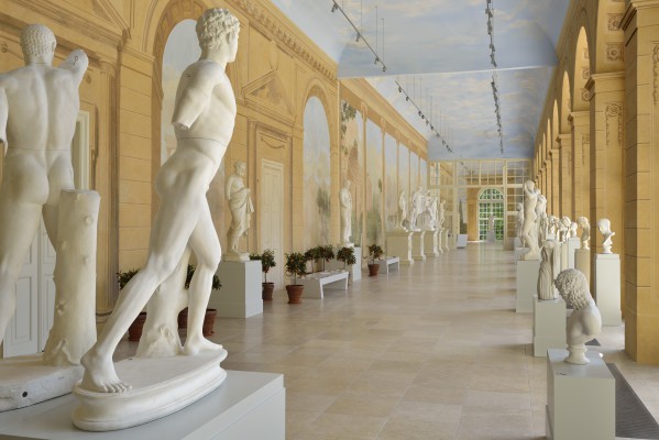 Widok ogólny na Galerię Rzeźby w Starej Oranżerii. Białe, marmurowe posągi stoją w rzędzie wzdłuż ściany tworzonej przez filary i wysokie okna oraz wzdłuż ściany ozdobionej malowidłami przedstawiającymi drzewa, góry i antyczne zabudowania.