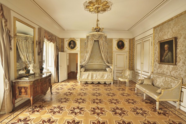 Sypialnia w Pałacu na Wyspie. Pośrodku stoi łóżko z baldachimem, po lewej stronie komoda, na którą wisi lustro, po prawej stronie stoi kanapa, nad którą wisi portret króla.