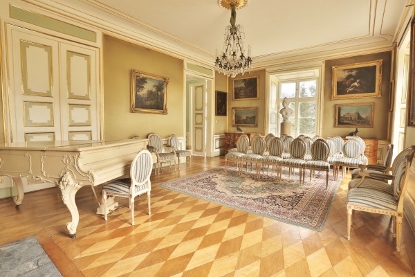 Salon w Pałacu Myślewickim, po prawej stronie stoi fortepian, na środku ustawione są krzesła w rzędach, krzesła stoją też pod ścianami po prawej i lewej stronie, ściany zdobią obrazy.