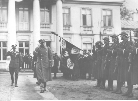 Archiwale zdjęcie przedstawiające żołnierzy stojących w szeregu i maszerujących przed nim dwóch dowódców.