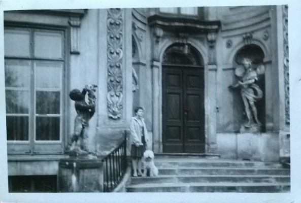Kobieta stojąca na schodach przed Pałacem Myślewickim, przy jej nogach siedzi pies.