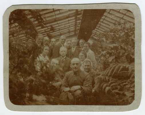 Archiwalna fotografia przedstawiająca grupę osób pozujących do zdjęcia w szklarni, w otoczeniu roślin.