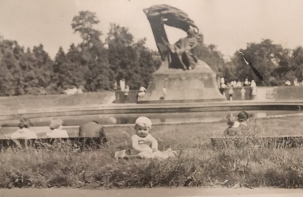 Dziecko siedzi na trawie, w tle widać Pomnik Fryderyka Chopina.