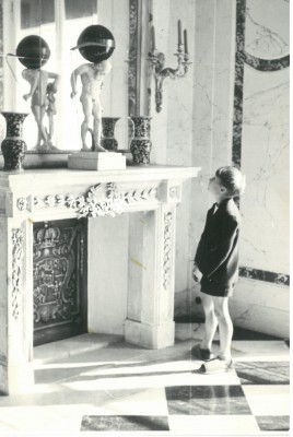 Dziecko stojące przed kominkiem w Pałacu na Wyspie. Na kominku stoi rzeźba przedstawiająca mężczyznę, trzymającego na barkach kulę.