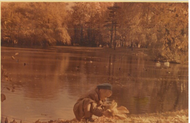 Dziecko siedzące nad brzegiem stawu, wokół którego rosną drzewa.