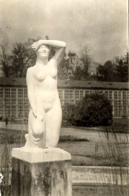 Rzeźba przedstawiająca nagą, klęczącą kobietę, rzeźba stoi na tle budynku. 