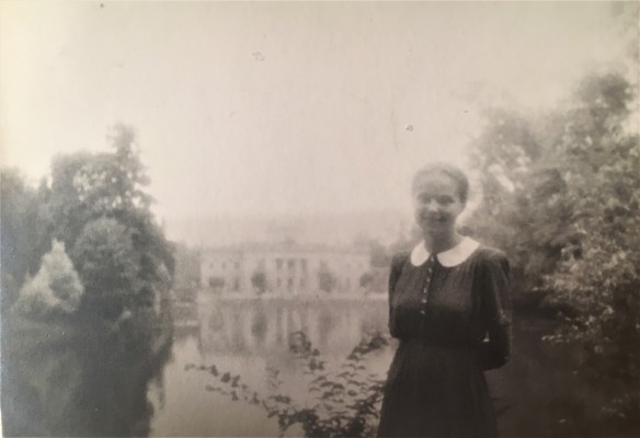 Kobieta stojąca w łazienkowskim ogrodzie, w tle widać Pałac na Wyspie otoczony wodą. 
