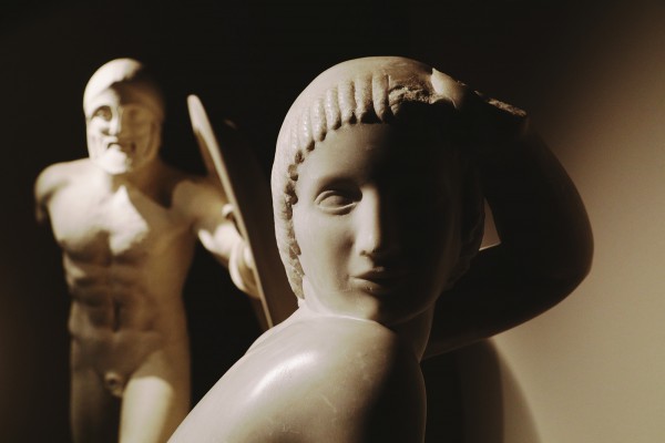 Rzeźba przedstawiająca kobietę, w tle, z tyłu, stoi rzeźba starożytnego wojownika. 