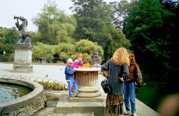 Dzieci przy fontannie w parku, obok stoją dwie kobiety, które rozmawiają.