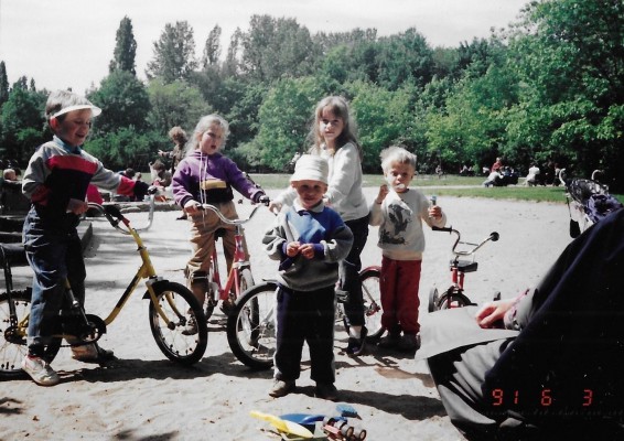 Grupa dzieci latem w parku, troje dzieci siedzi na rowerach.