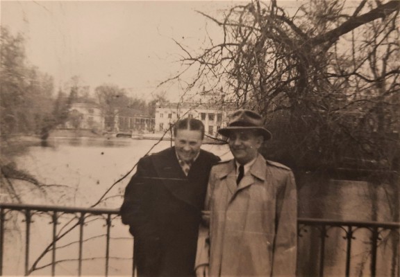 Dwaj mężczyźni stoją na moście przy barierce na tle wody i Pałacu na Wyspie w Łazienkach Królewskich.