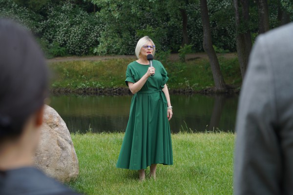 Kobieta w długiej zielonej sukience stoi na trawniku z mikrofonem w ręku. 