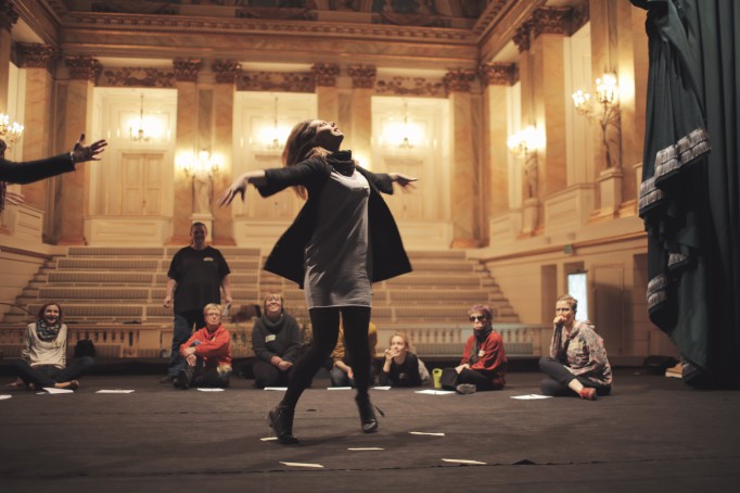 Scena w teatrze, na której siedzą ludzie, jedna z kobiet tańczy, unoszcząc ręce.