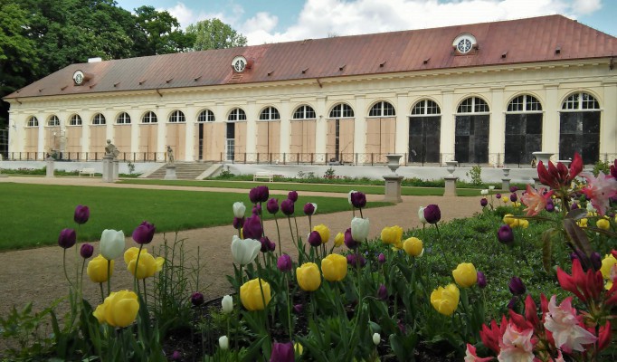 Budynek Starej Oranżerii w Łazienkach Królewskch, widać fragment ogrodu, w którym kwitną żółte, białe i bordowe kwiaty.